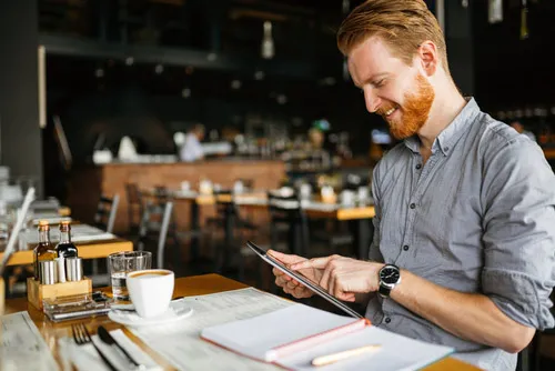 A smiling man in a casual shirt sits in an empty restaurant with notebooks and smiles into a tablet