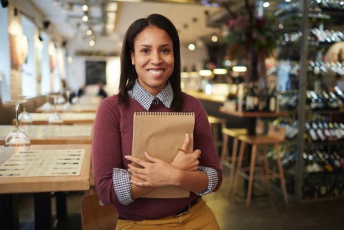 A restaurant manager stands in front of an empty restaurant interior while holding a large notepad