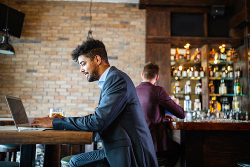 A suited man sits at a high table in a restaurant and looks into a laptop