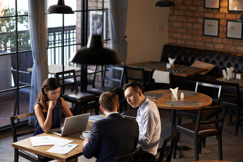 Three professionals sit discussing in an empty restaurant with papers and a laptop on the table